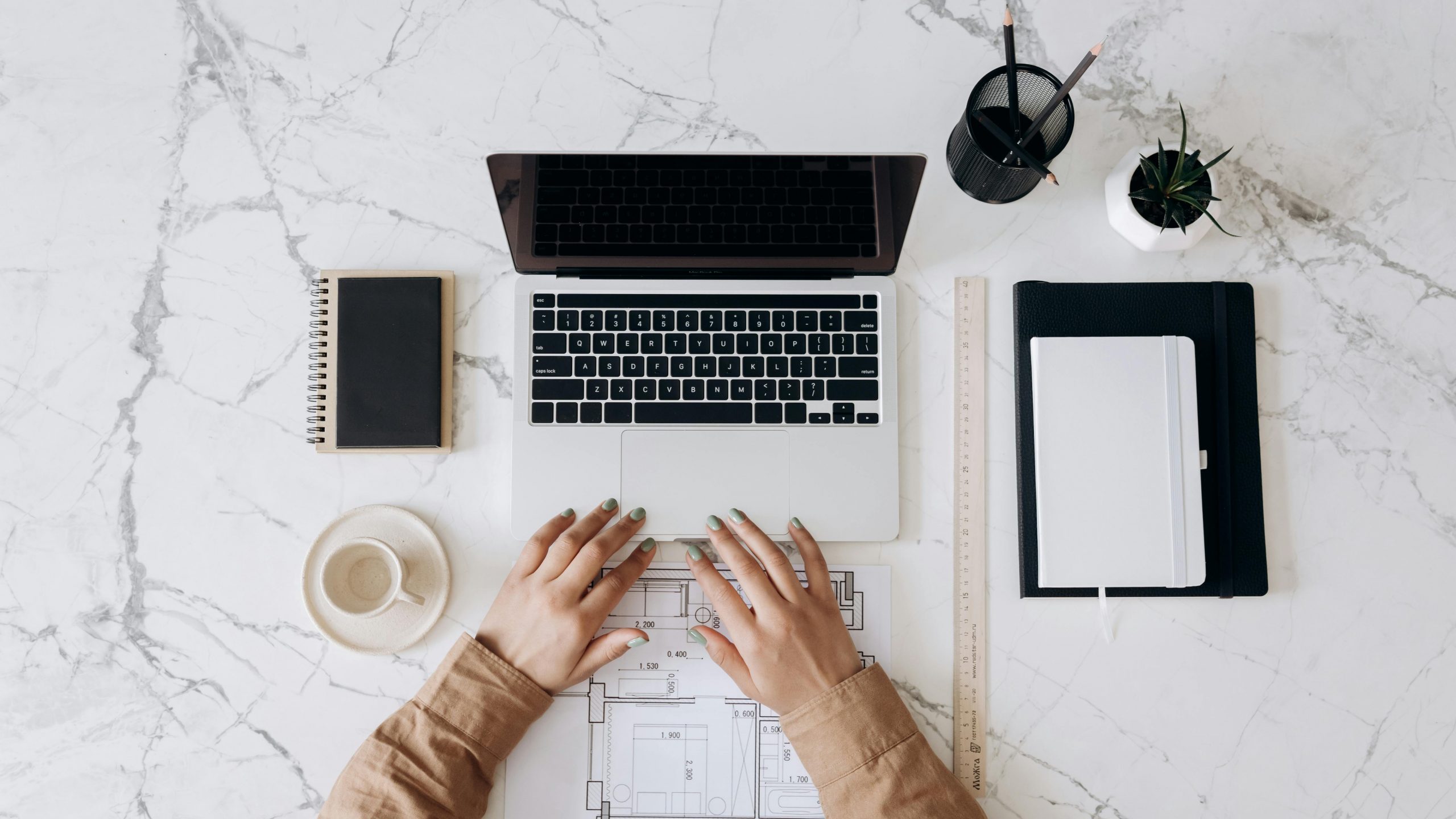 Top view of a stylish home office desk with a laptop, planner, and coffee cup, showing hands on a blueprint.